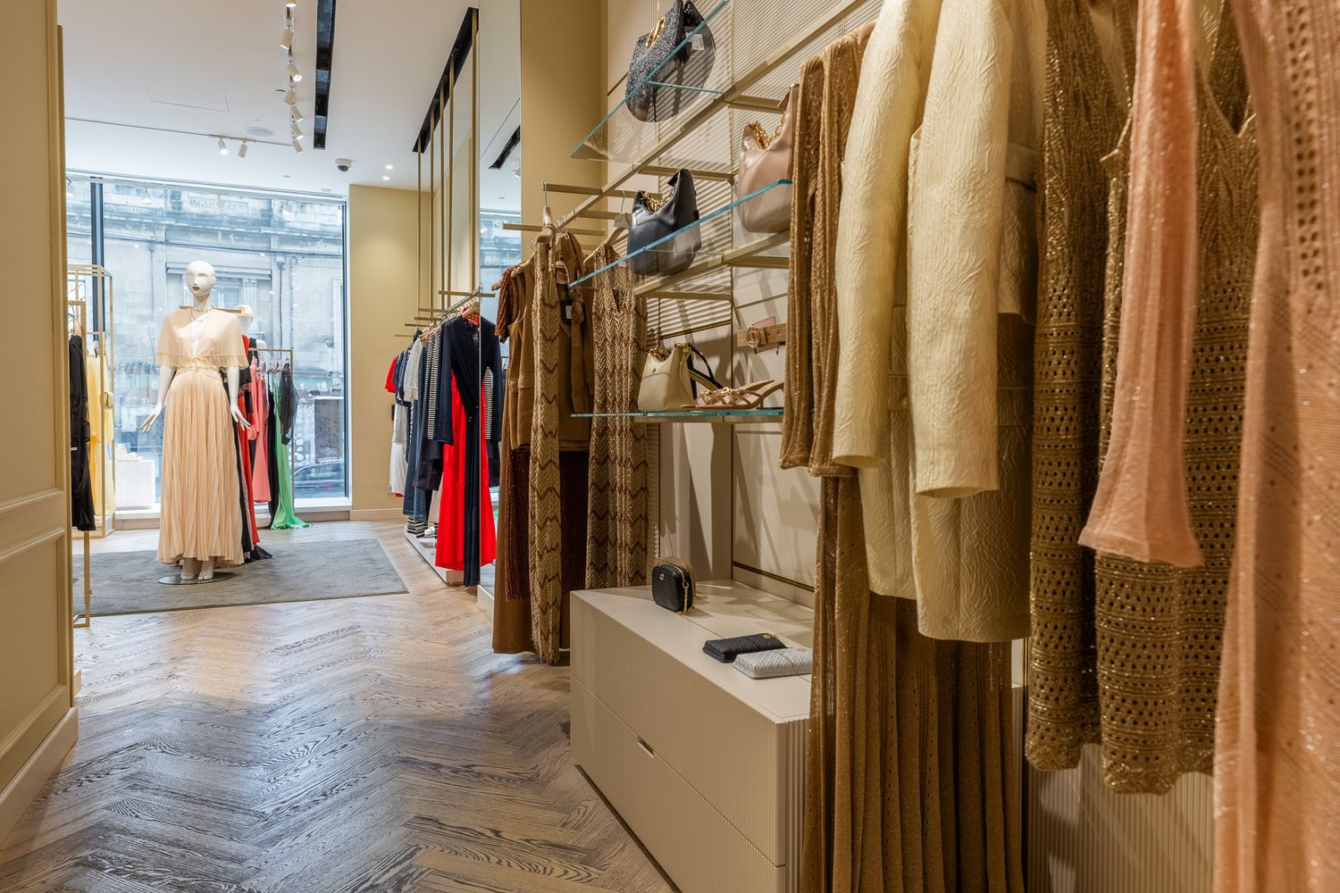 Chic boutique interior with herringbone floors, mannequin, and colorful garments hanging on gold racks.