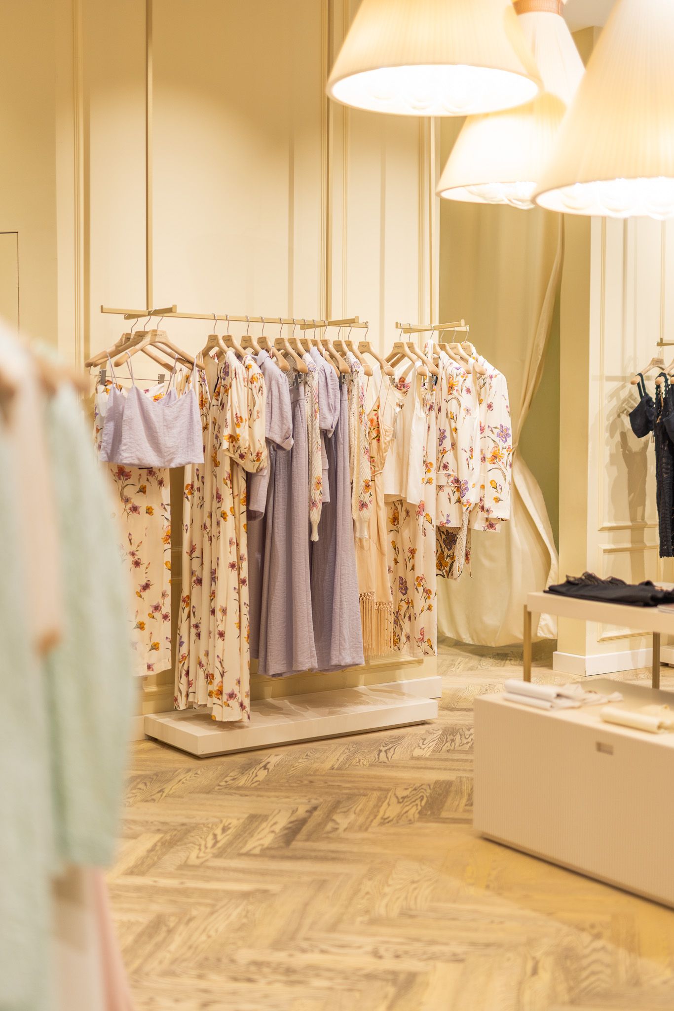 Bright boutique interior with floral and pastel dresses on wooden racks under warm pendant lights.