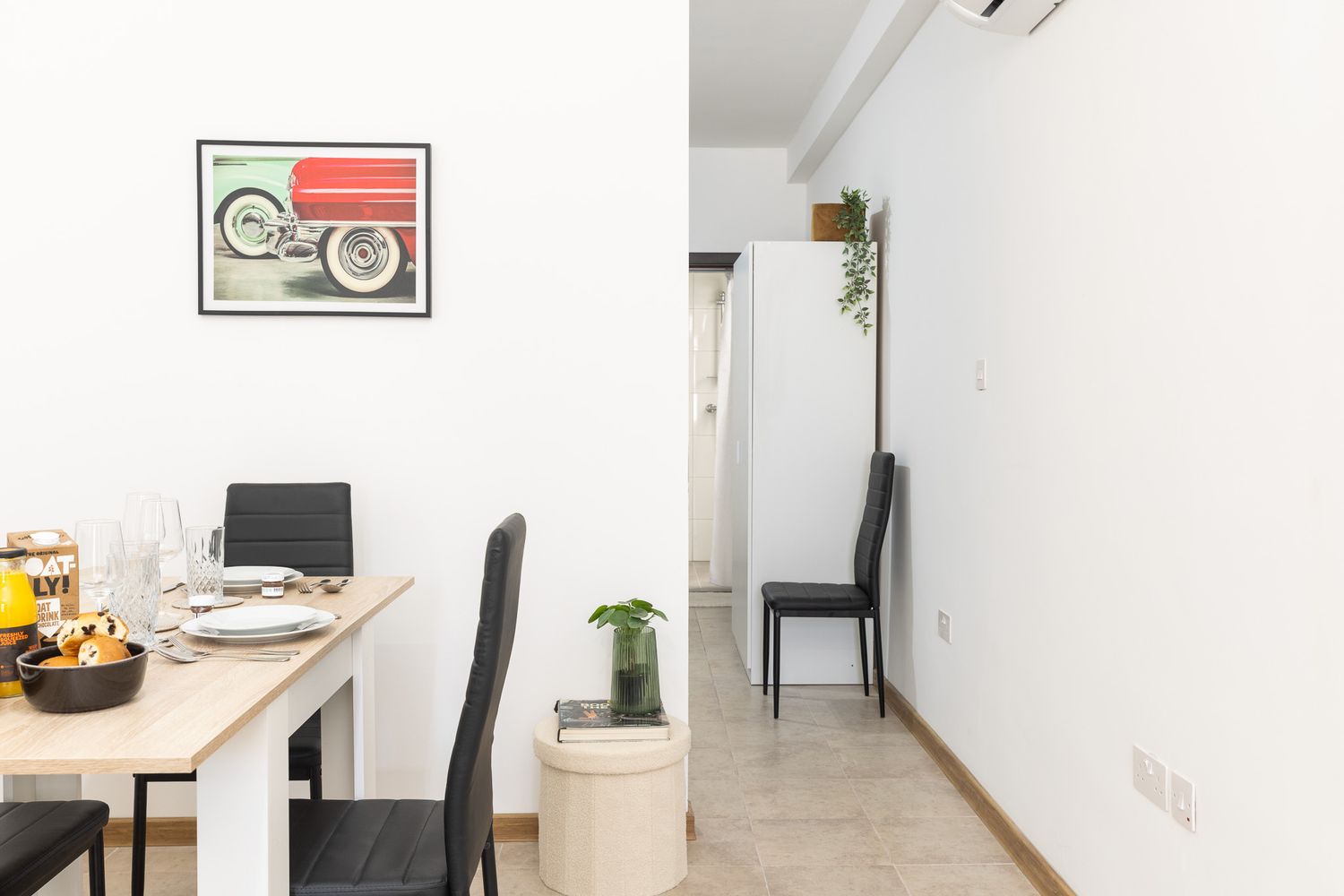 Compact dining area with wooden table, black chairs, vintage car artwork, and open hallway view.
