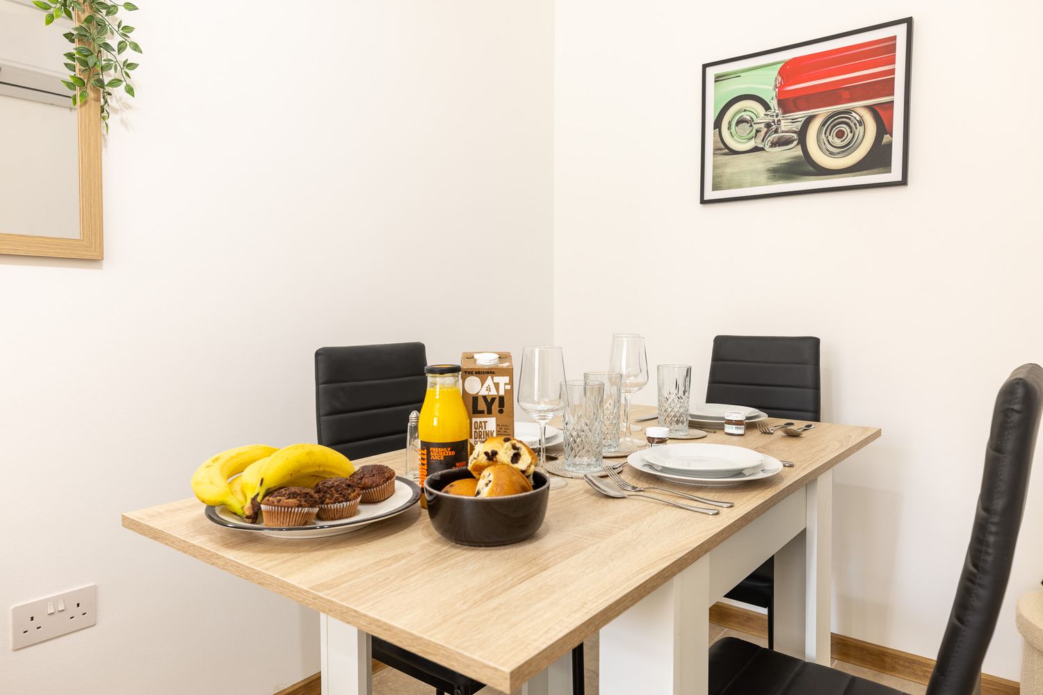 Dining table set with fresh fruit, orange juice, and tableware beneath a vintage red car artwork.