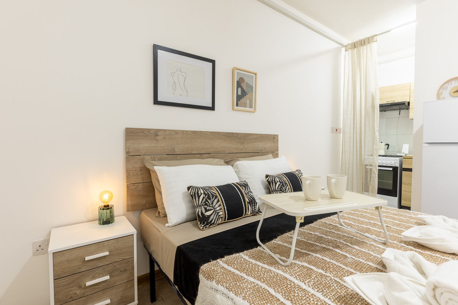 Cozy studio apartment bedroom with wooden headboard, patterned pillows, bedside lamp, and glimpse of kitchen area.