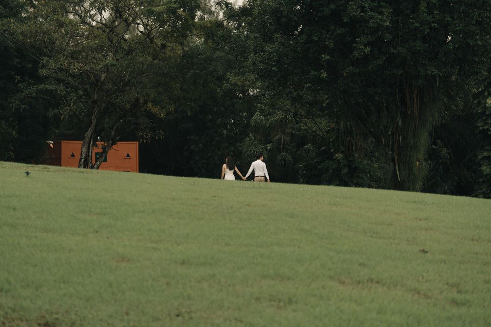 Couple holding hands walking across lush green meadow with rustic wooden structure and dense trees in background.