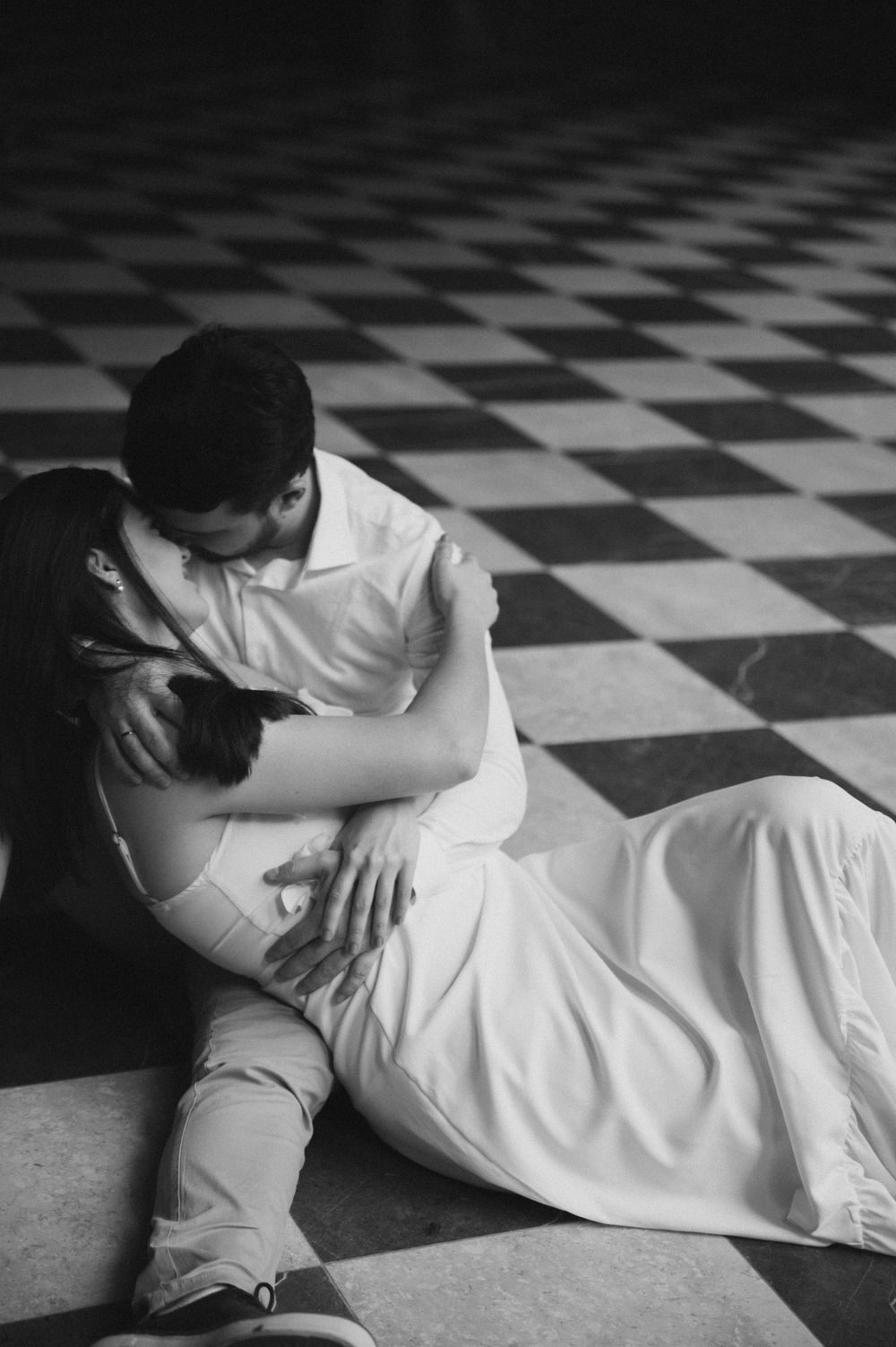 Black and white photo of a couple kissing on a checkered floor, the woman in a flowing white dress.