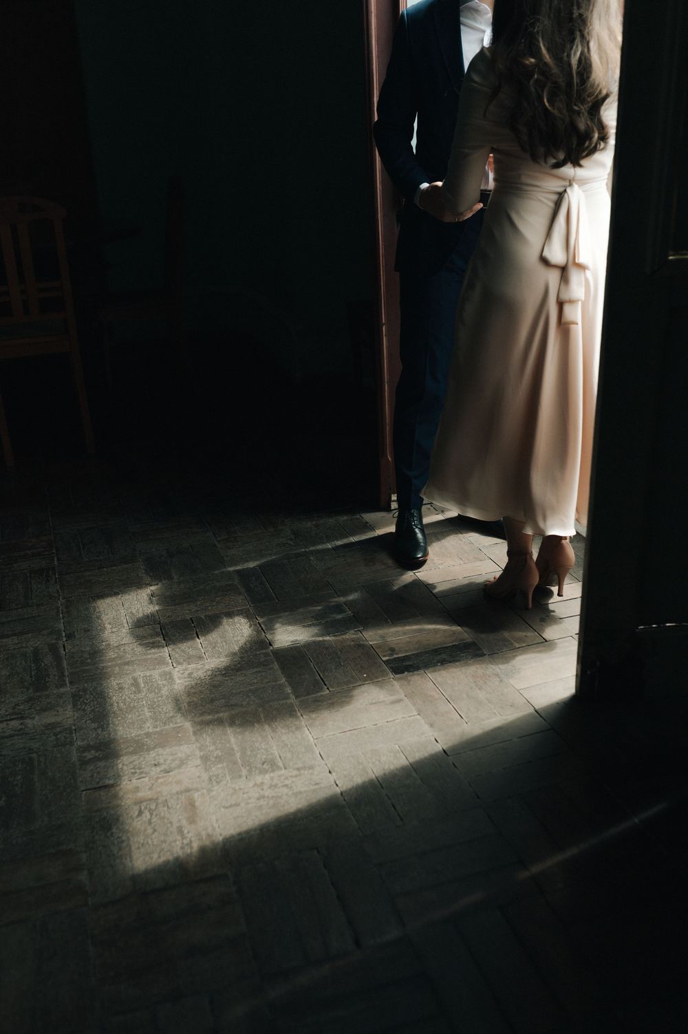 Couple standing in a doorway casting shadows on tiled floor, woman in elegant cream dress and man in dark suit.