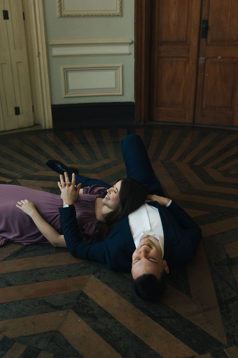 Romantic couple lying on herringbone parquet floor in elegant venue, woman in purple gown, man in navy suit.