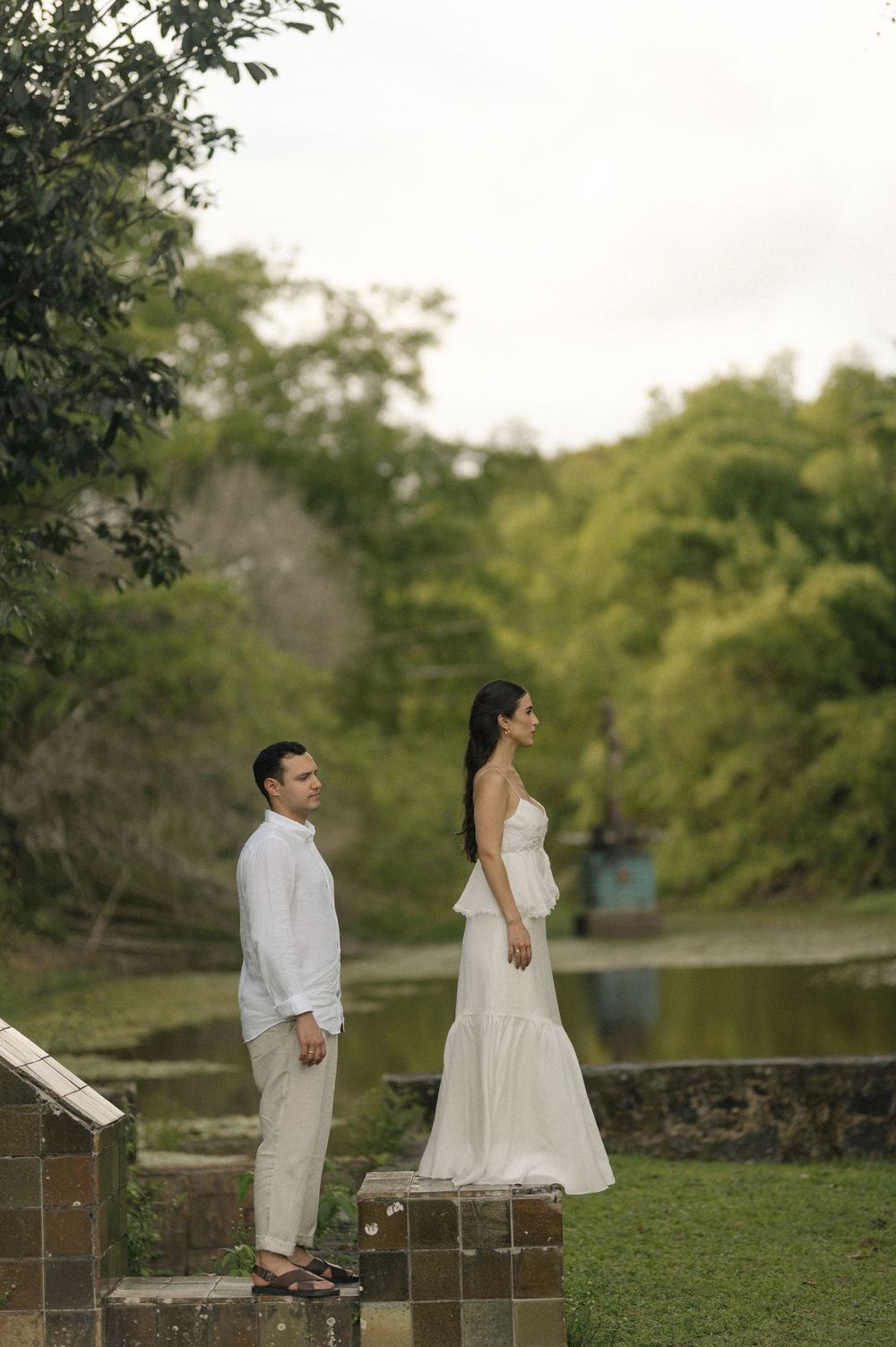 Couple in white attire standing on stone pillars surrounded by lush greenery near a serene pond.