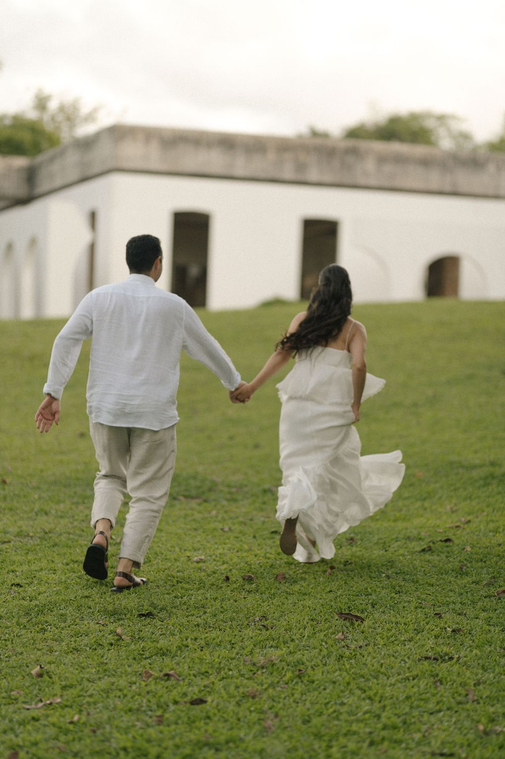 Couple in white wedding attire holding hands while walking across green grass toward a white building.