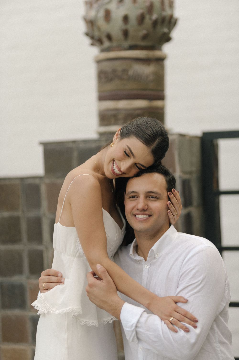 A smiling couple in white outfits embrace warmly outdoors near a stone column backdrop.