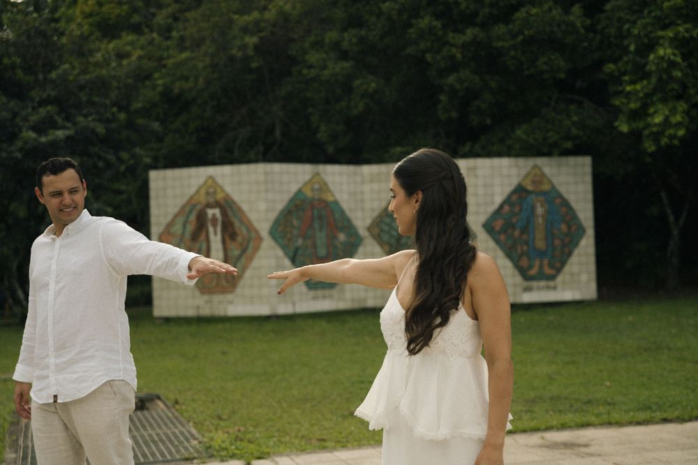 A couple in white attire pointing at each other in a park with colorful mosaic artwork in the background.