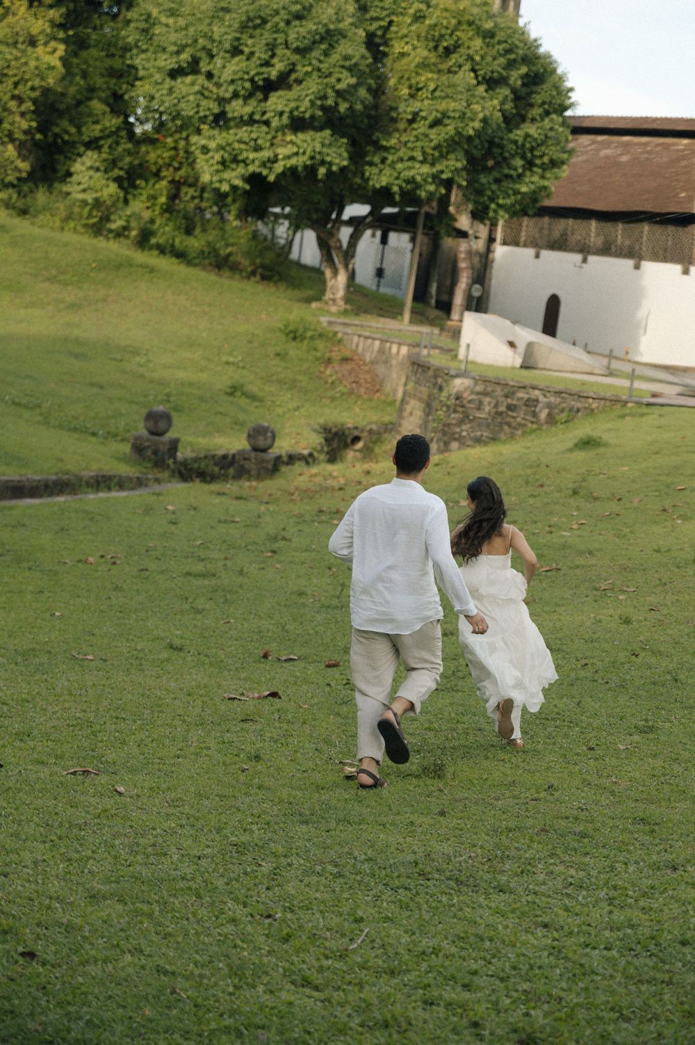 Couple in white outfits holding hands running up a green grassy hill toward trees and a white building.