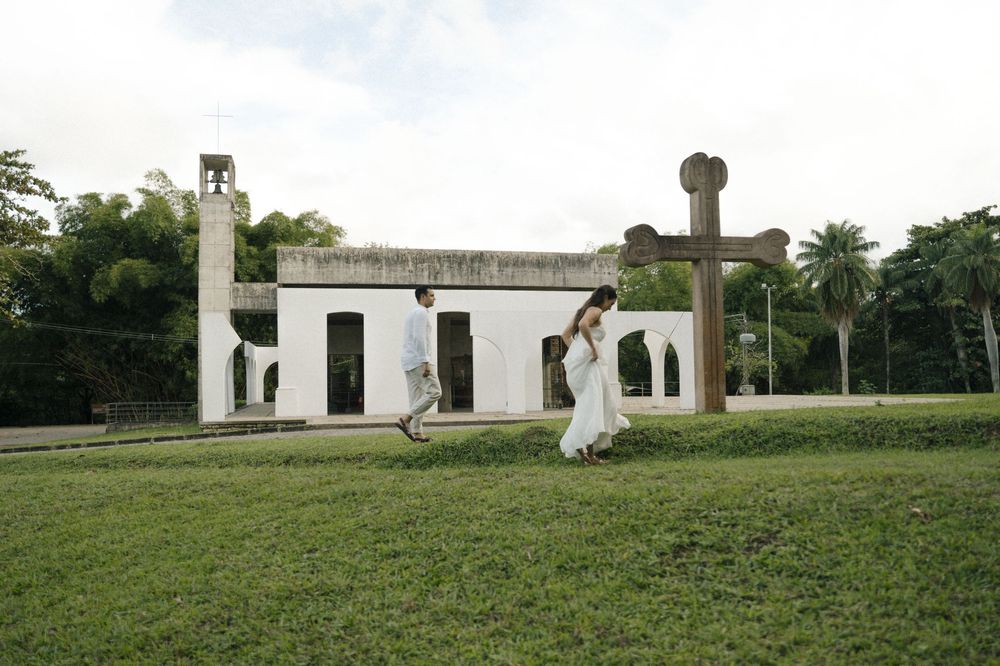 A woman in a white dress walks past a large wooden cross on a green lawn outside a historic white church.