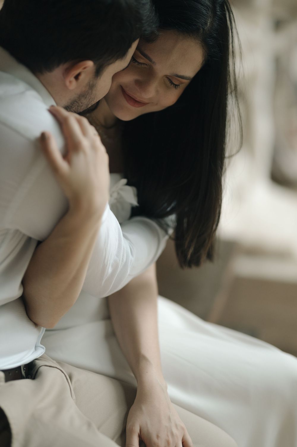 A smiling dark-haired woman in a white dress embraces a man in a white shirt during a romantic couples photo session.