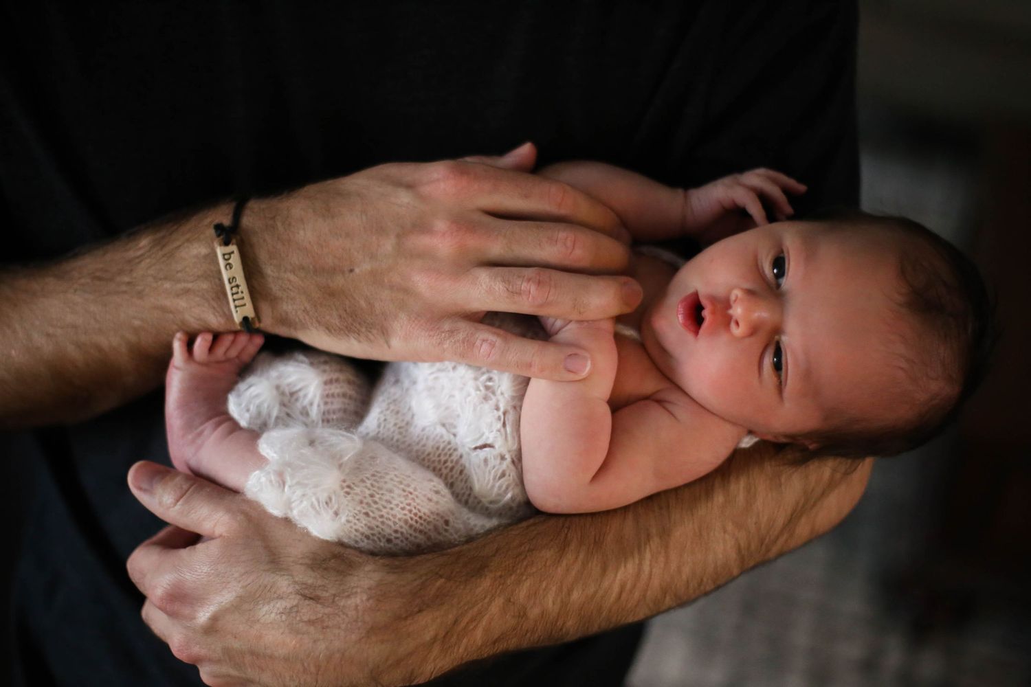 Newborn baby girl in stretched out in Dad's arms with bracelet that reads "be still".