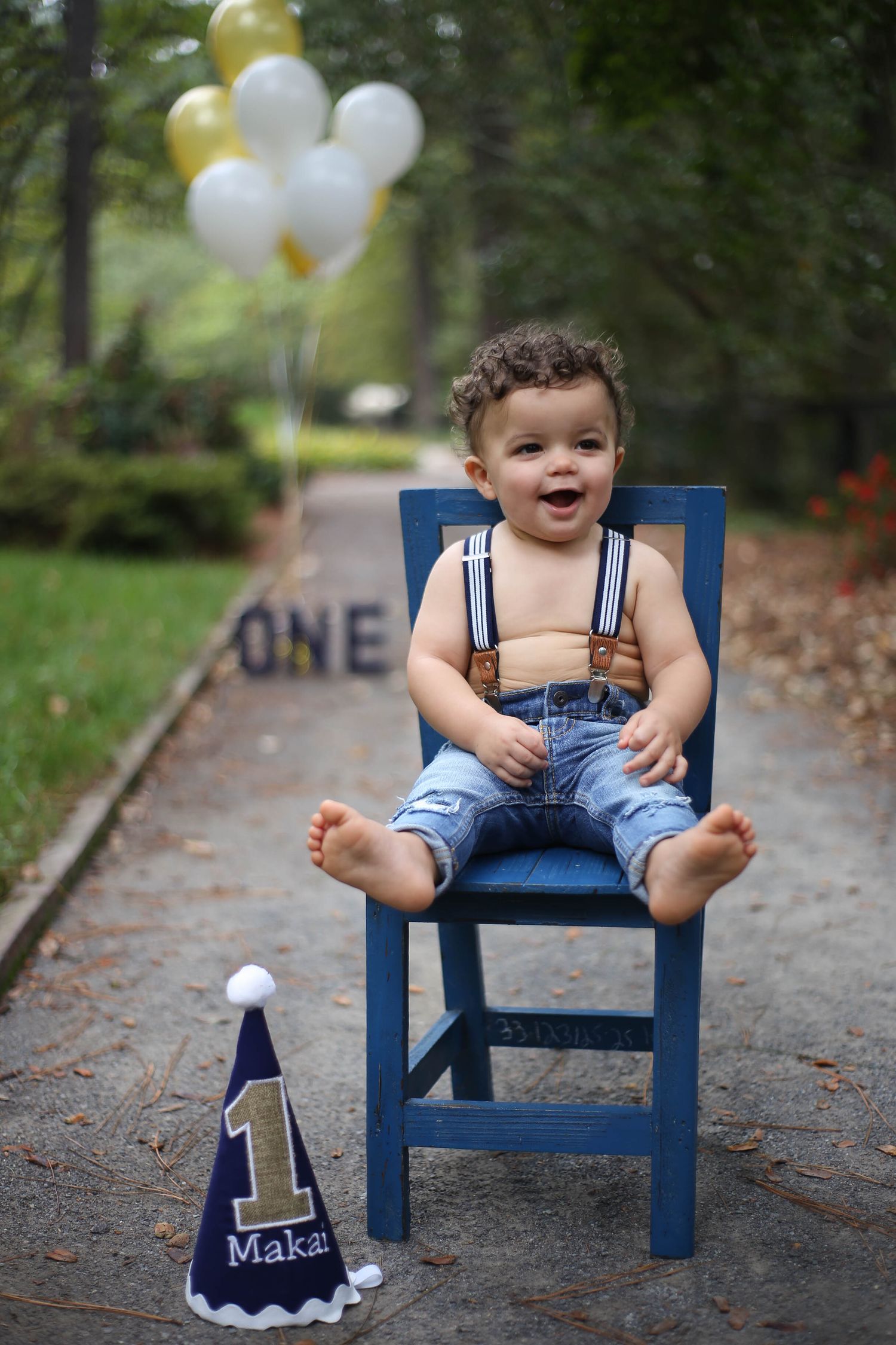 Happy chubby baby boy in suspenders sits in a chair in the gardens.  Celebrating his first birthday.