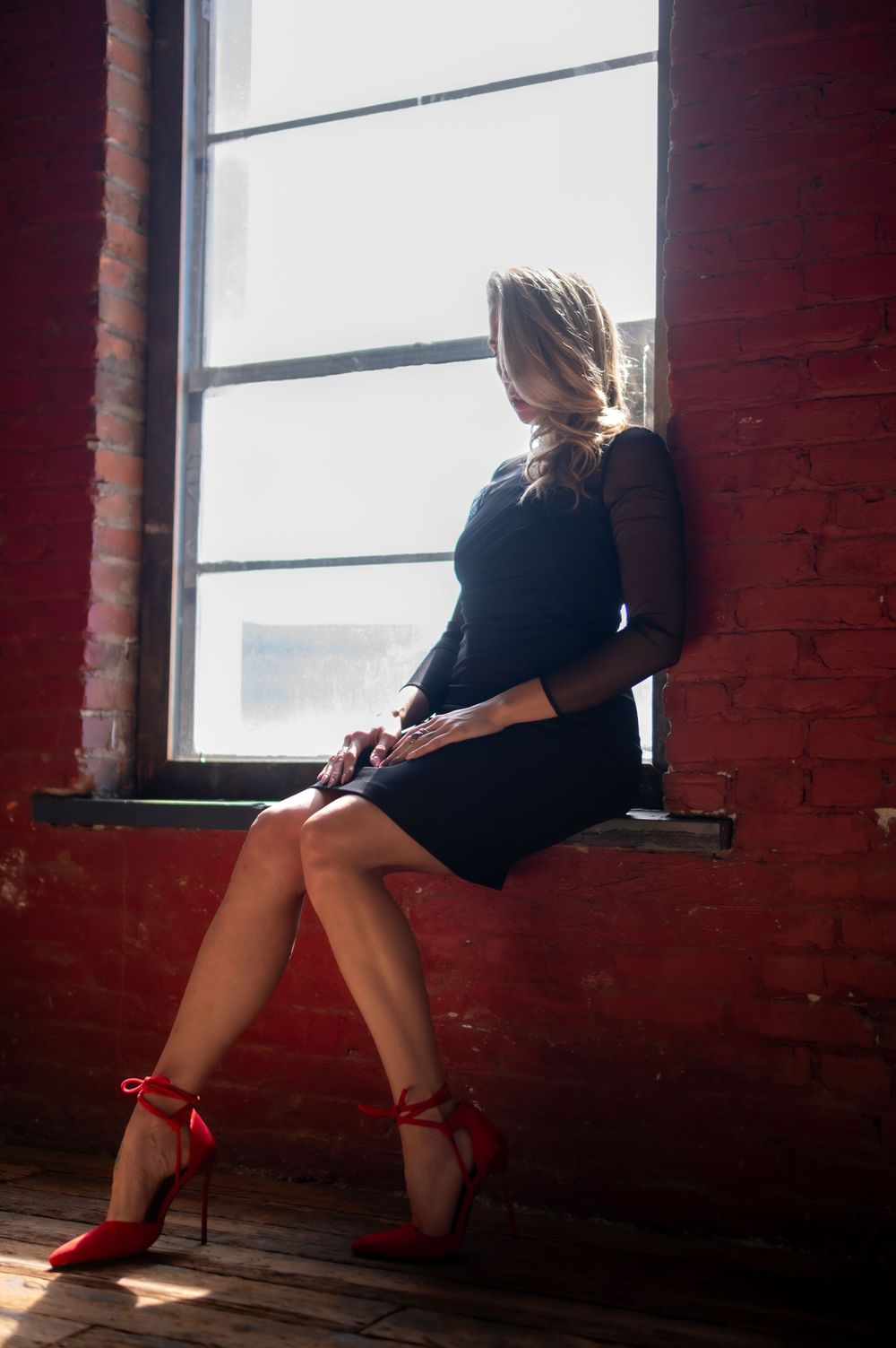 Woman in black dress with red heels sitting on windowsill against red brick wall.