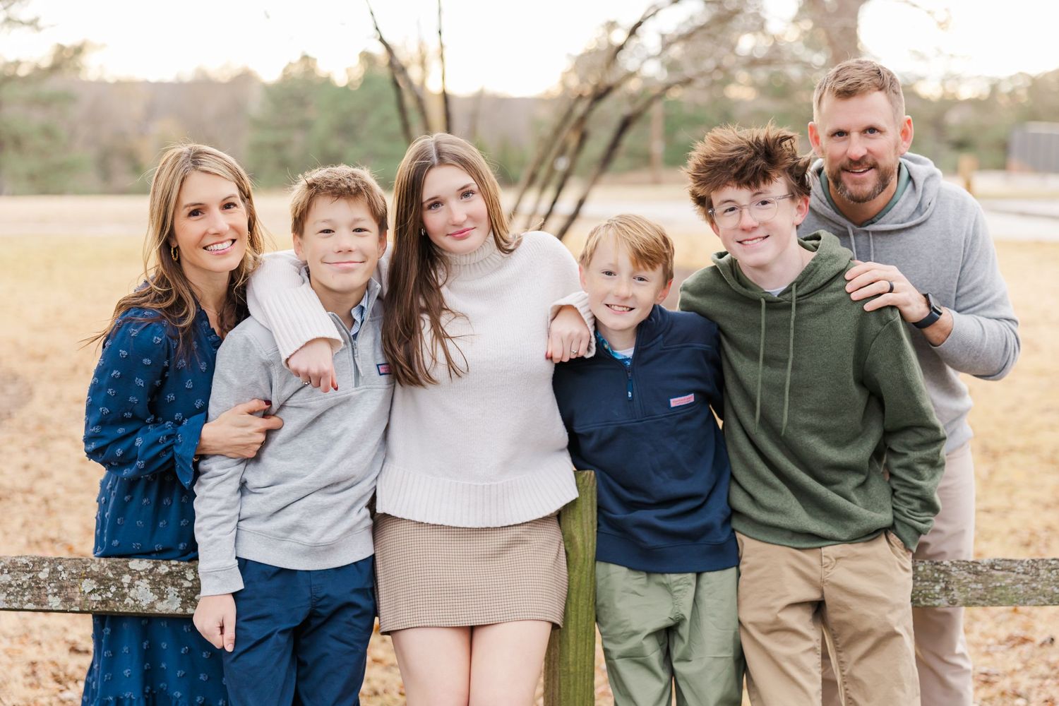 Family of six stands together outdoors in casual wear leaning on a wooden fence during autumn.