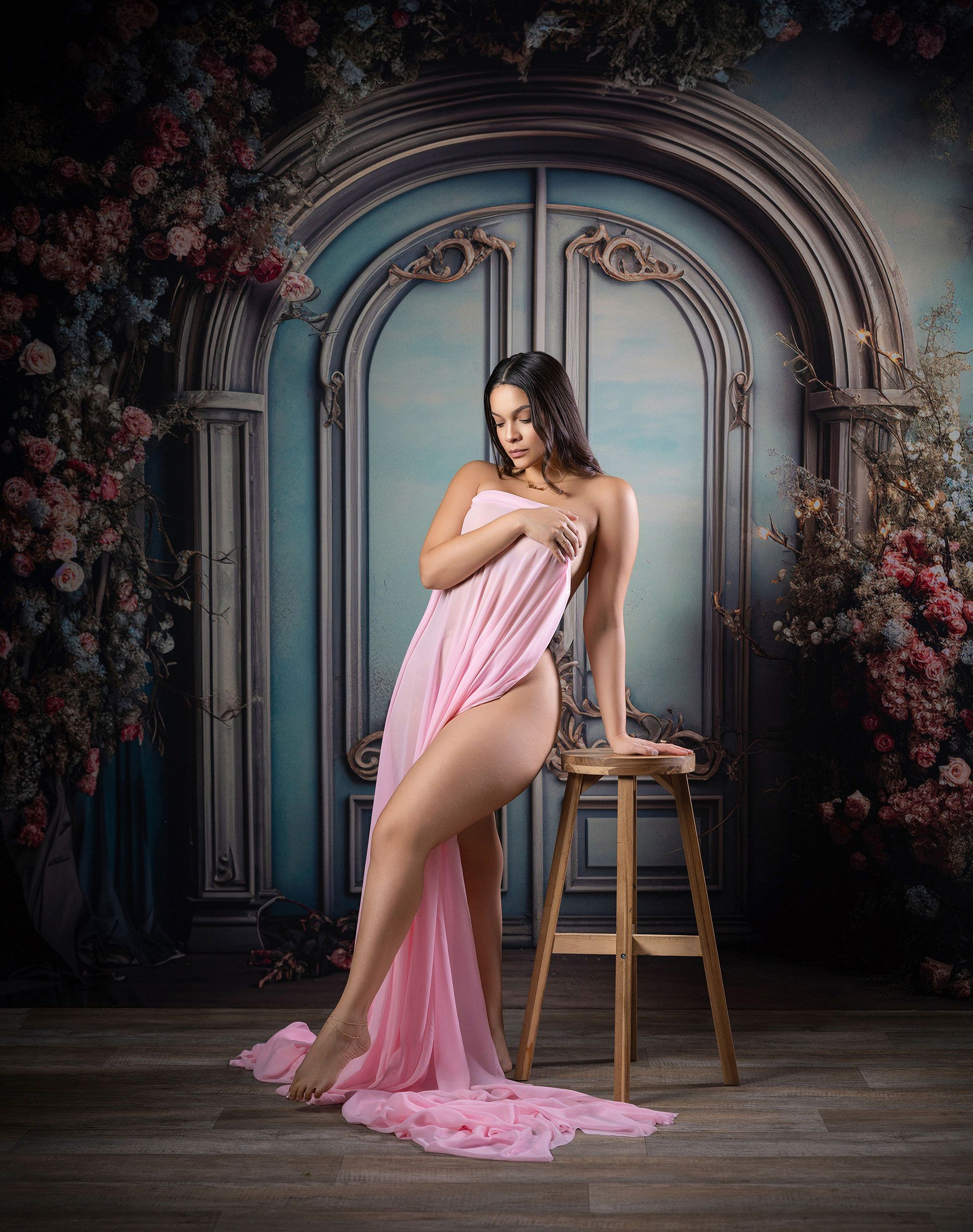 Artistic boudoir photo with flowing pink fabric in front of ornate blue doors with floral arch and vintage stool setup.