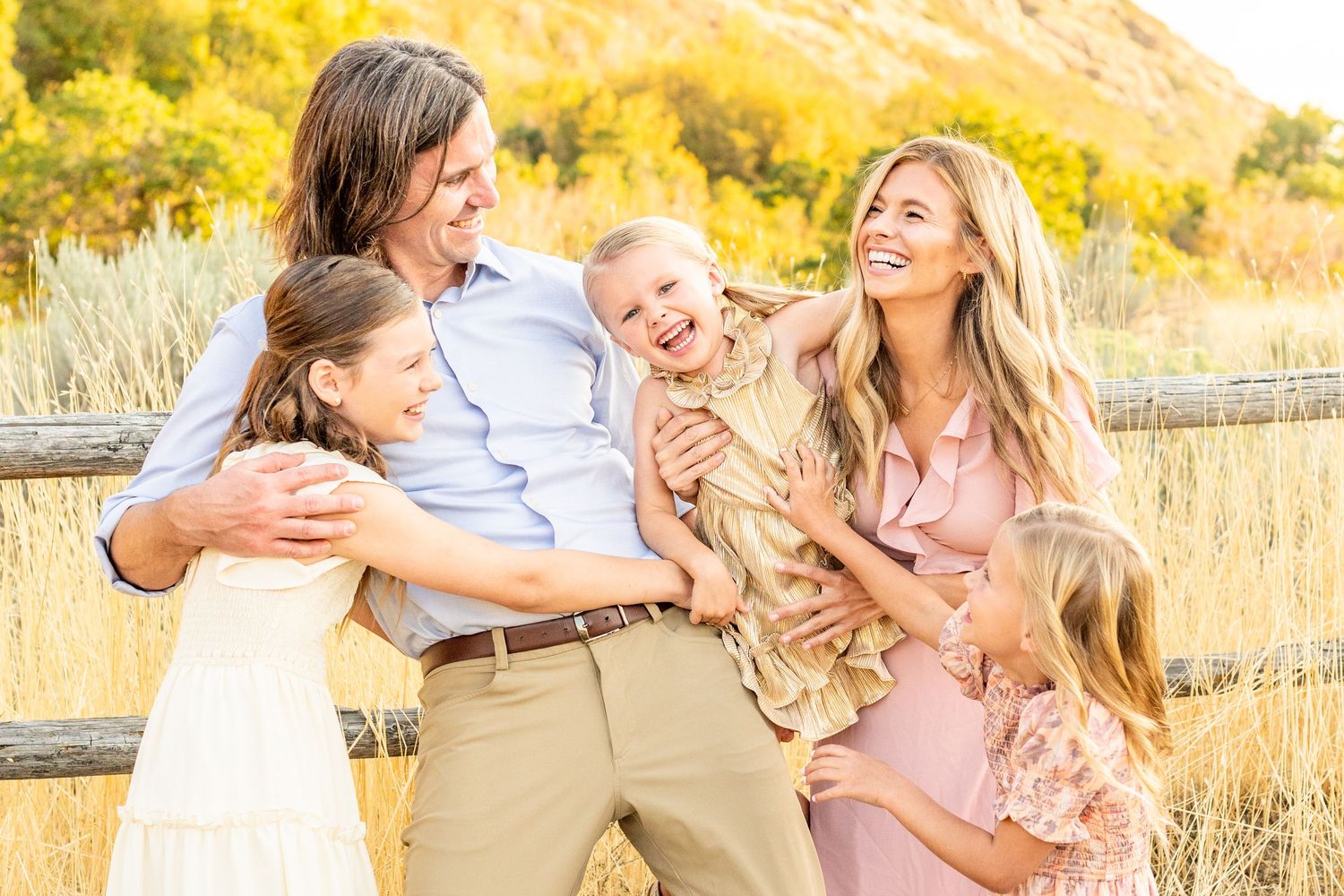 Candid family moment in a sun-lit field with mountain backdrop wearing coordinated pink and white outfits at Hidden Valley Park, Utah.