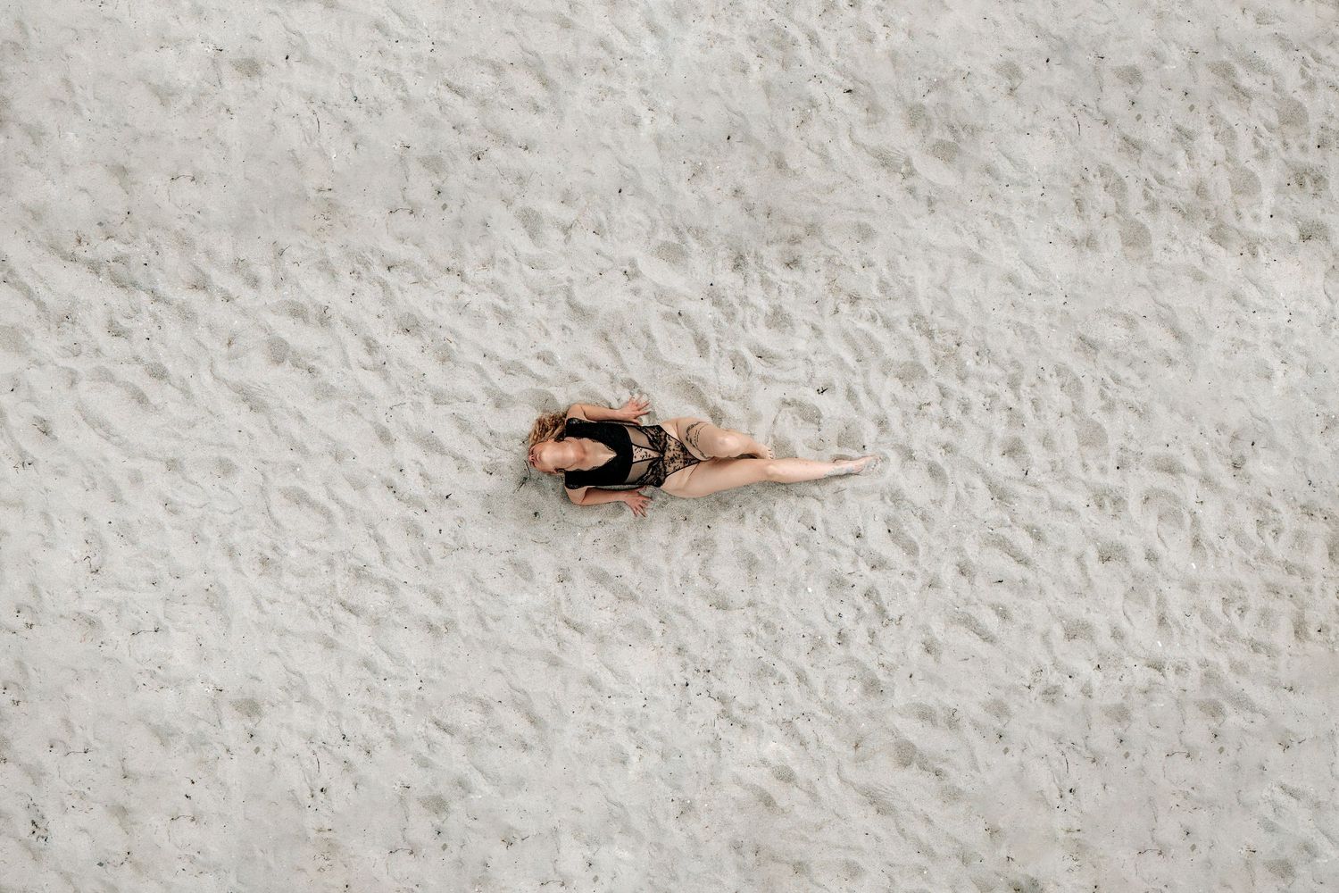 Dronefoto af en person i badedragt liggende på sandet i et kunstnerisk boudoir-portræt