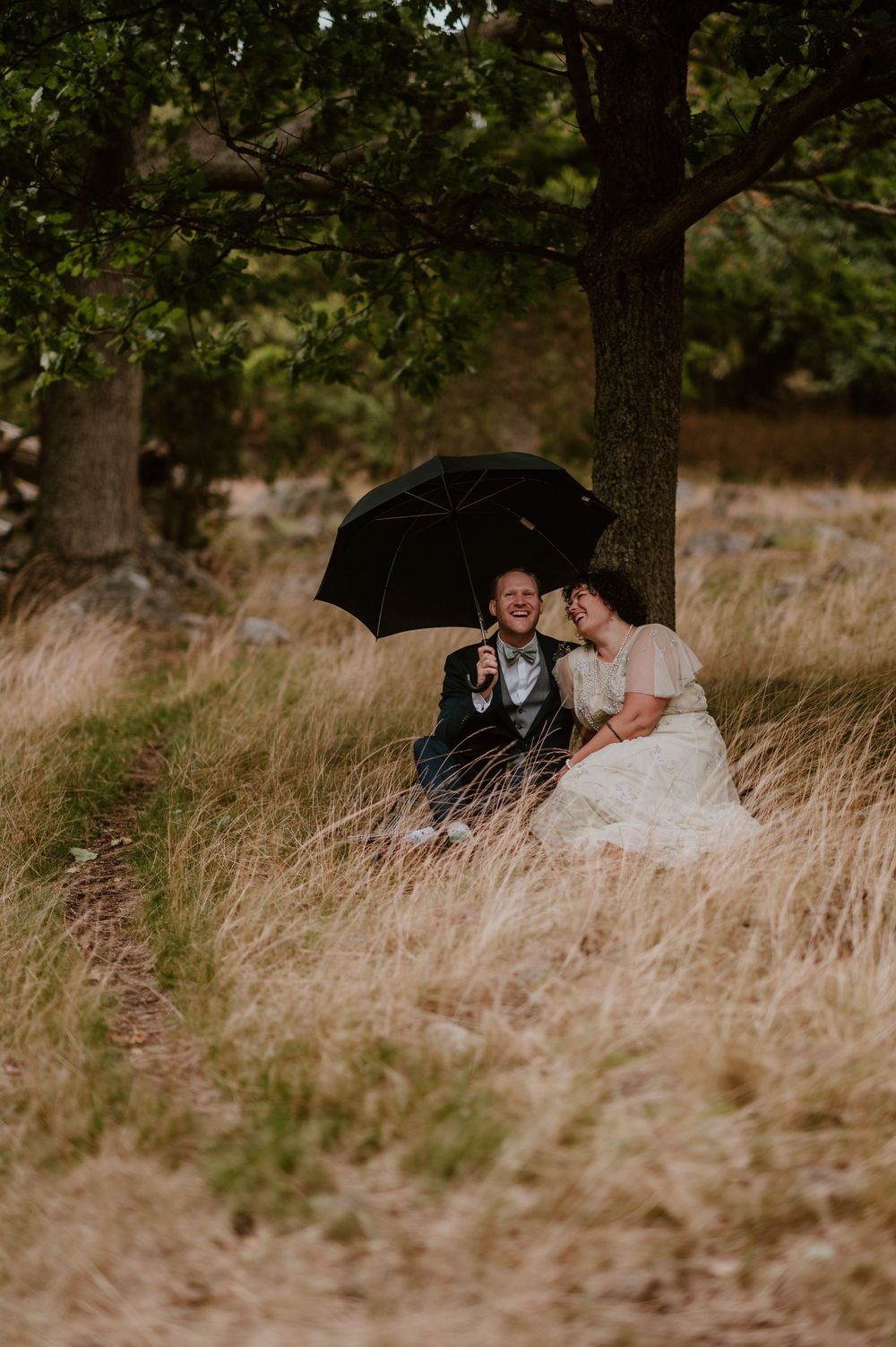 A romantic couple sits together in tall golden grass beneath shady trees during a wedding photoshoot.