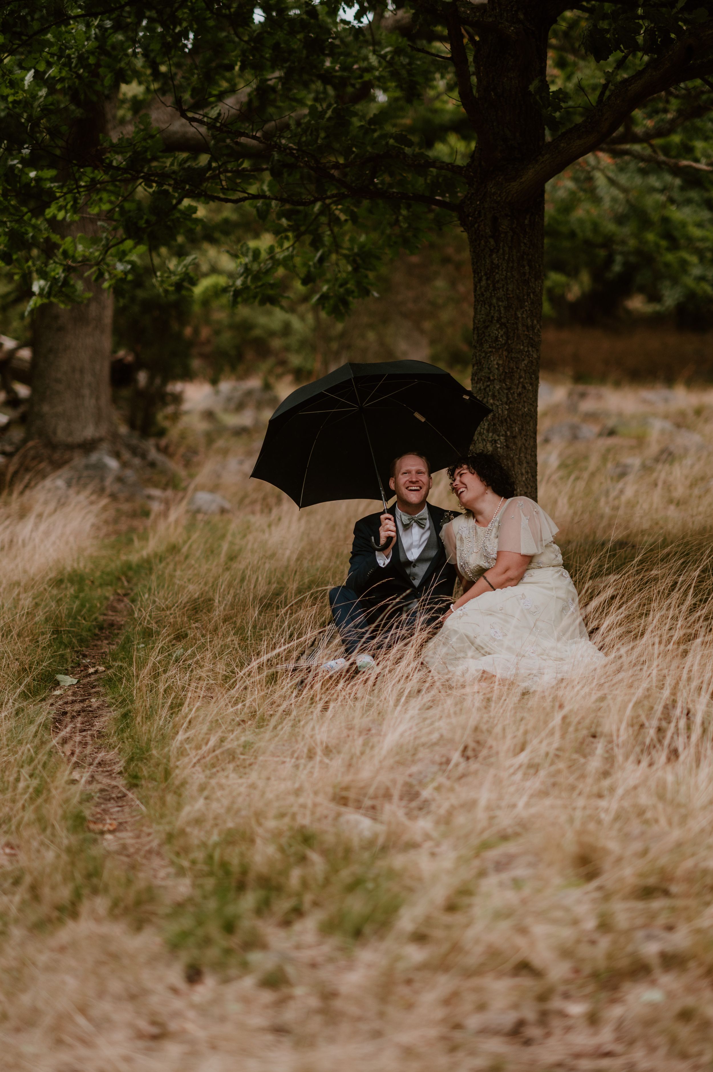 A romantic couple sits together in tall golden grass beneath shady trees during a wedding photoshoot.
