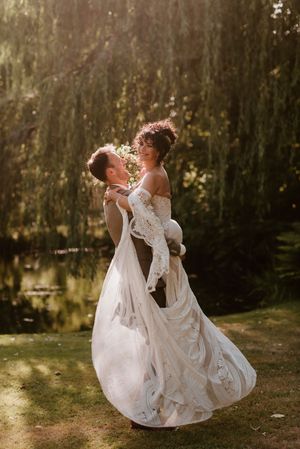 A couple in white wedding attire dances joyfully in a sunlit garden setting with willow trees in the background.