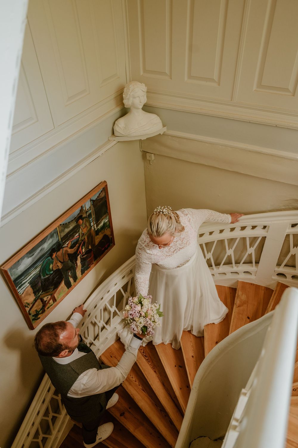 A bride in a white wedding dress ascends a grand staircase in an elegant interior with white walls and wooden steps.