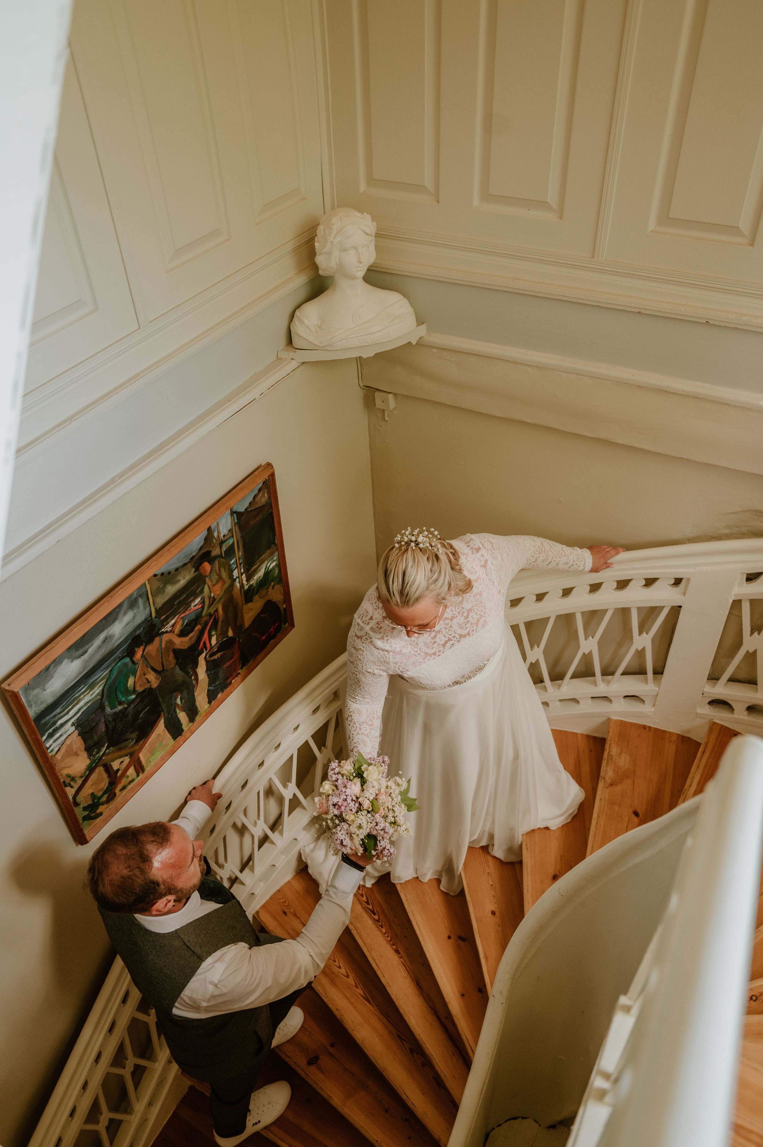 A bride in a white wedding dress ascends a grand staircase in an elegant interior with white walls and wooden steps.