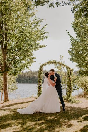 A couple shares a romantic moment under a floral arch by a lake on their wedding day with trees framing the scene.