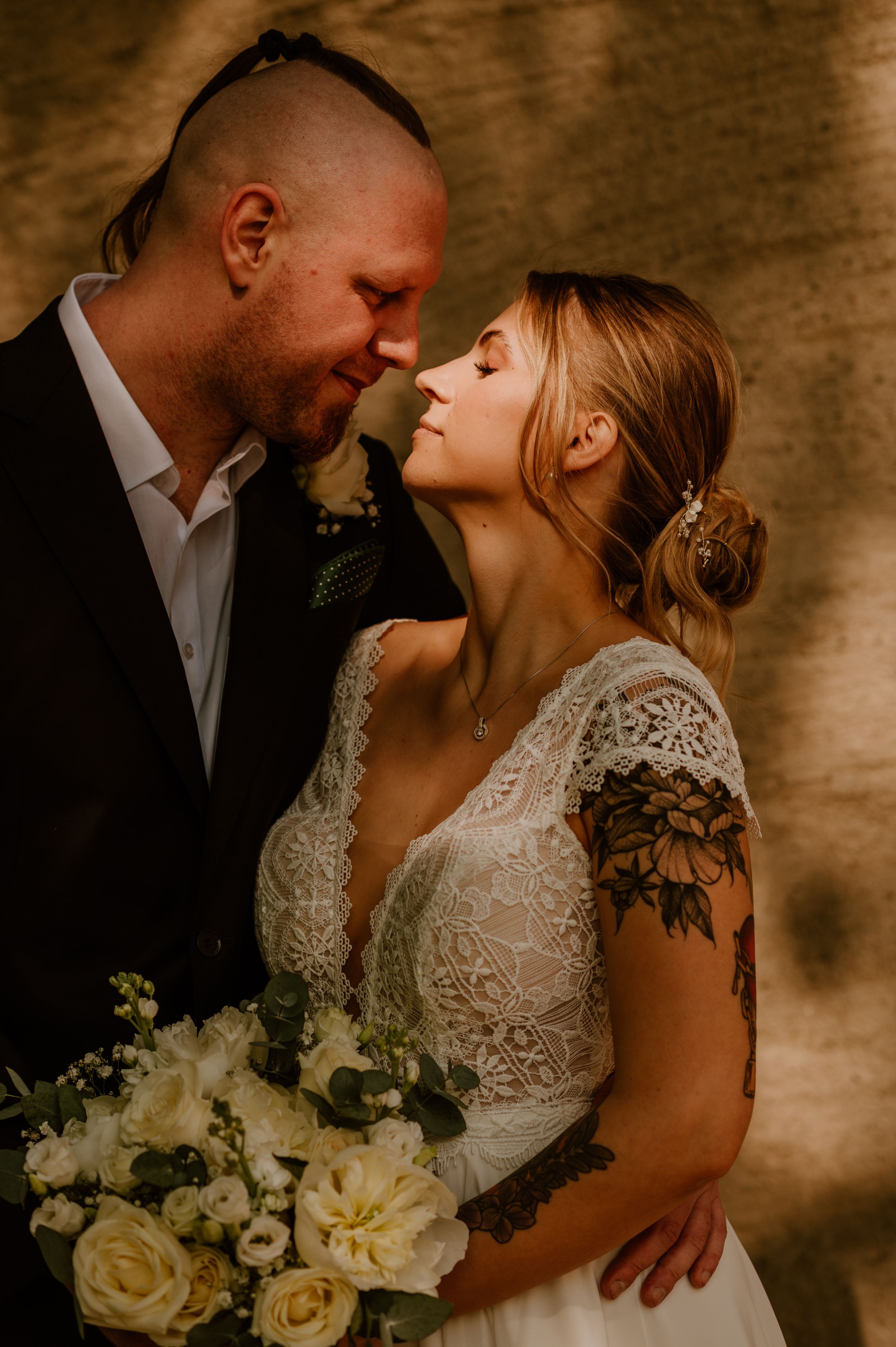 Romantic wedding couple embraces in soft lighting with white rose bouquet against textured brown backdrop.