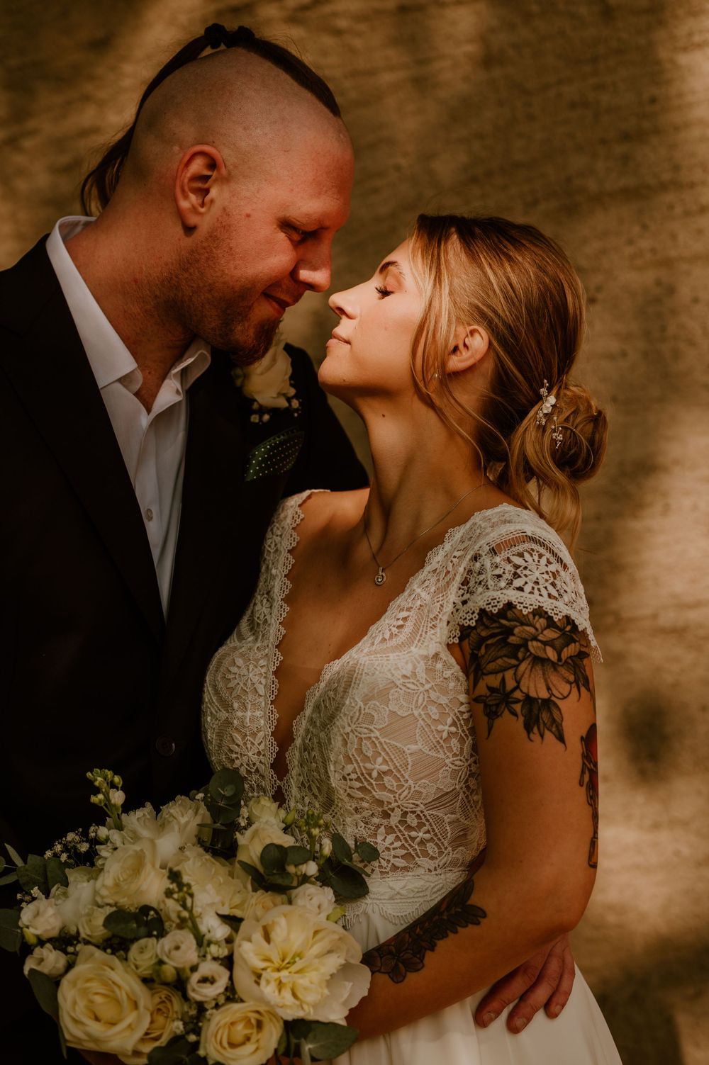 Romantic wedding couple embraces in soft lighting with white rose bouquet against textured brown backdrop.