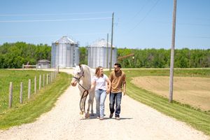 A couple walks a white horse down a rural gravel path with grain silos and green trees in the background.