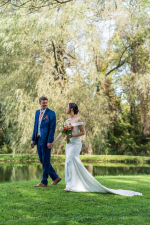 Newlyweds walk hand in hand by a pond under weeping willow trees in a romantic setting.