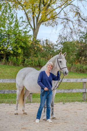 Woman in blue sweater and jeans smiling beside a white horse in a sandy paddock with autumn trees.