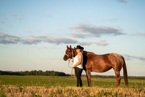 Woman in black cowboy hat standing beside a chestnut horse in a green field under a blue sky.