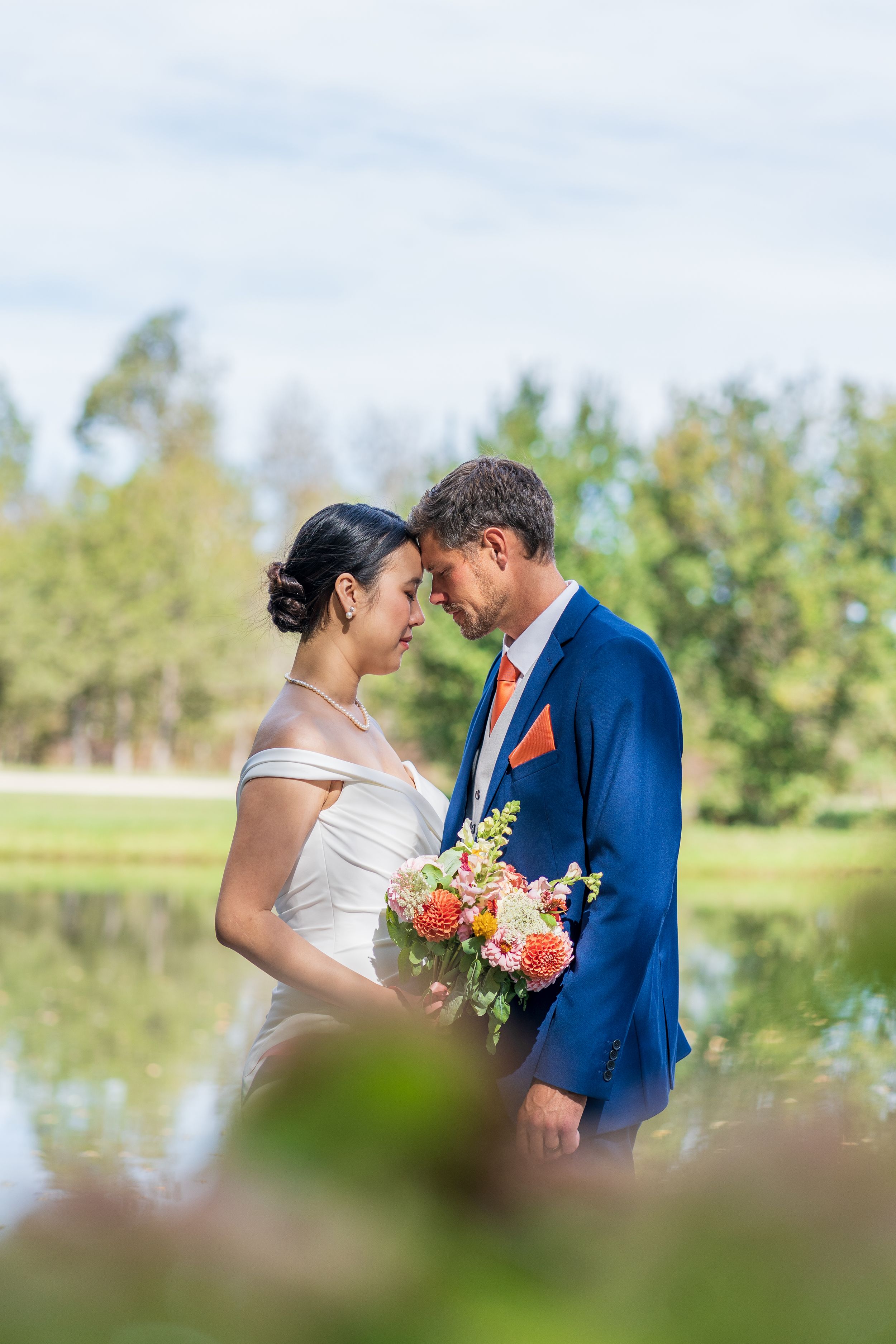 A romantic wedding couple sharing an intimate moment by a pond with a colorful bouquet.
