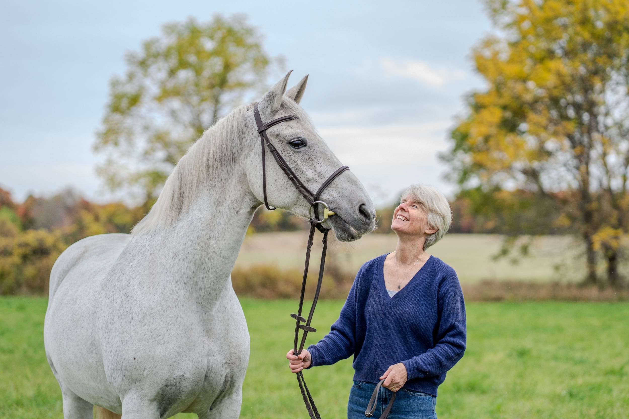 Elderly woman in blue sweater smiling up at a white horse in a green field with autumn trees.