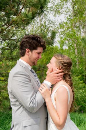 A groom in a gray suit tenderly holds his bride's face as they share a romantic moment outdoors among green trees.