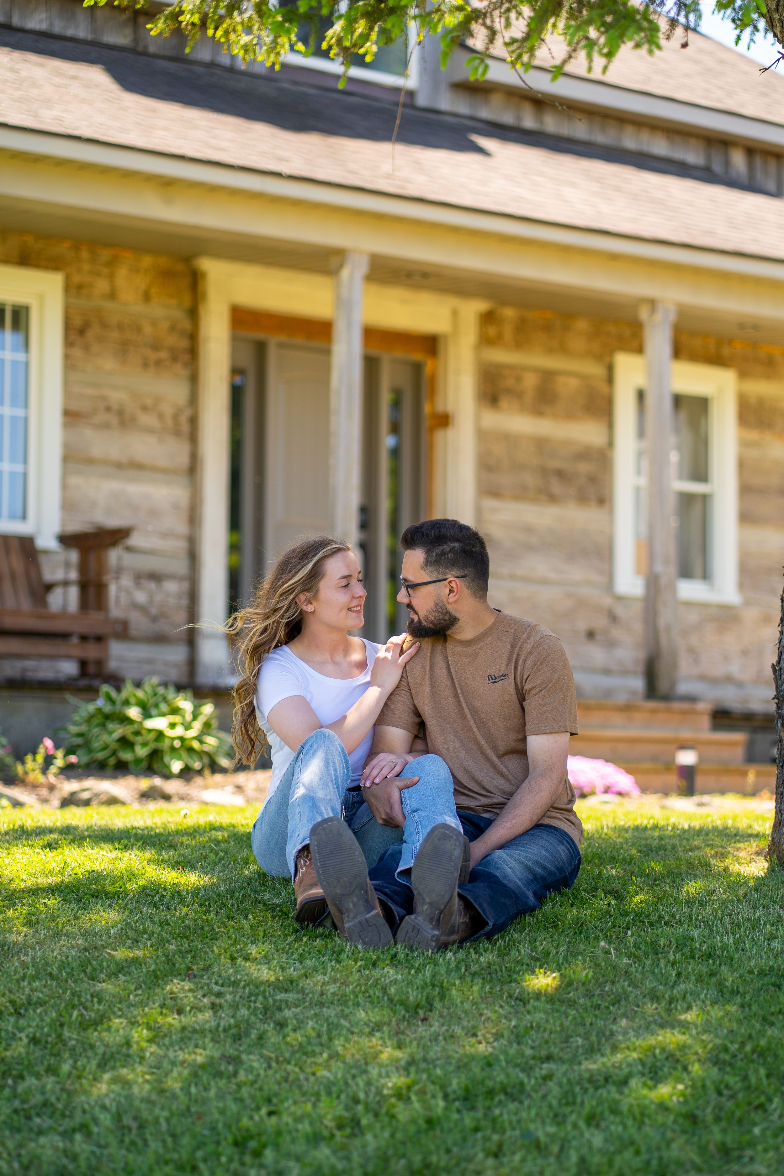 Couple sitting on green grass in front of a rustic stone house, smiling at each other on a sunny day.