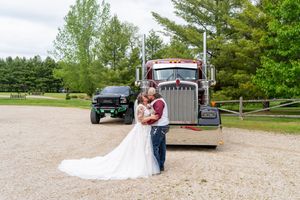 Bride and groom embracing in front of a Kenworth semi-truck on a gravel lot surrounded by trees.