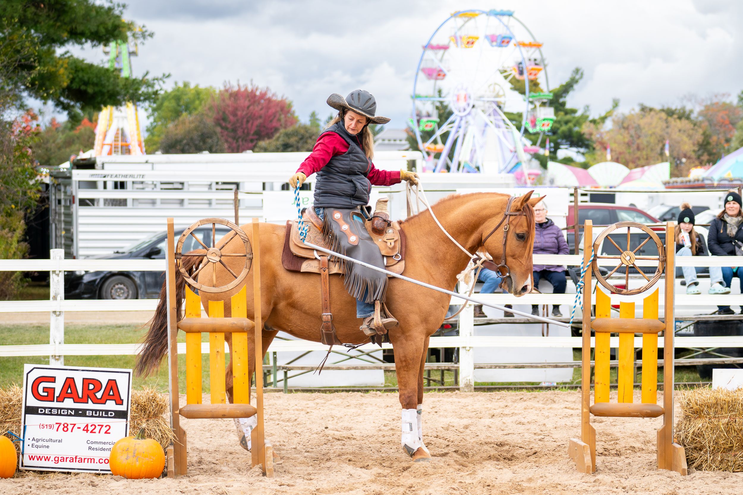 Equestrian navigates a gate obstacle at a fall competition with a ferris wheel in background.