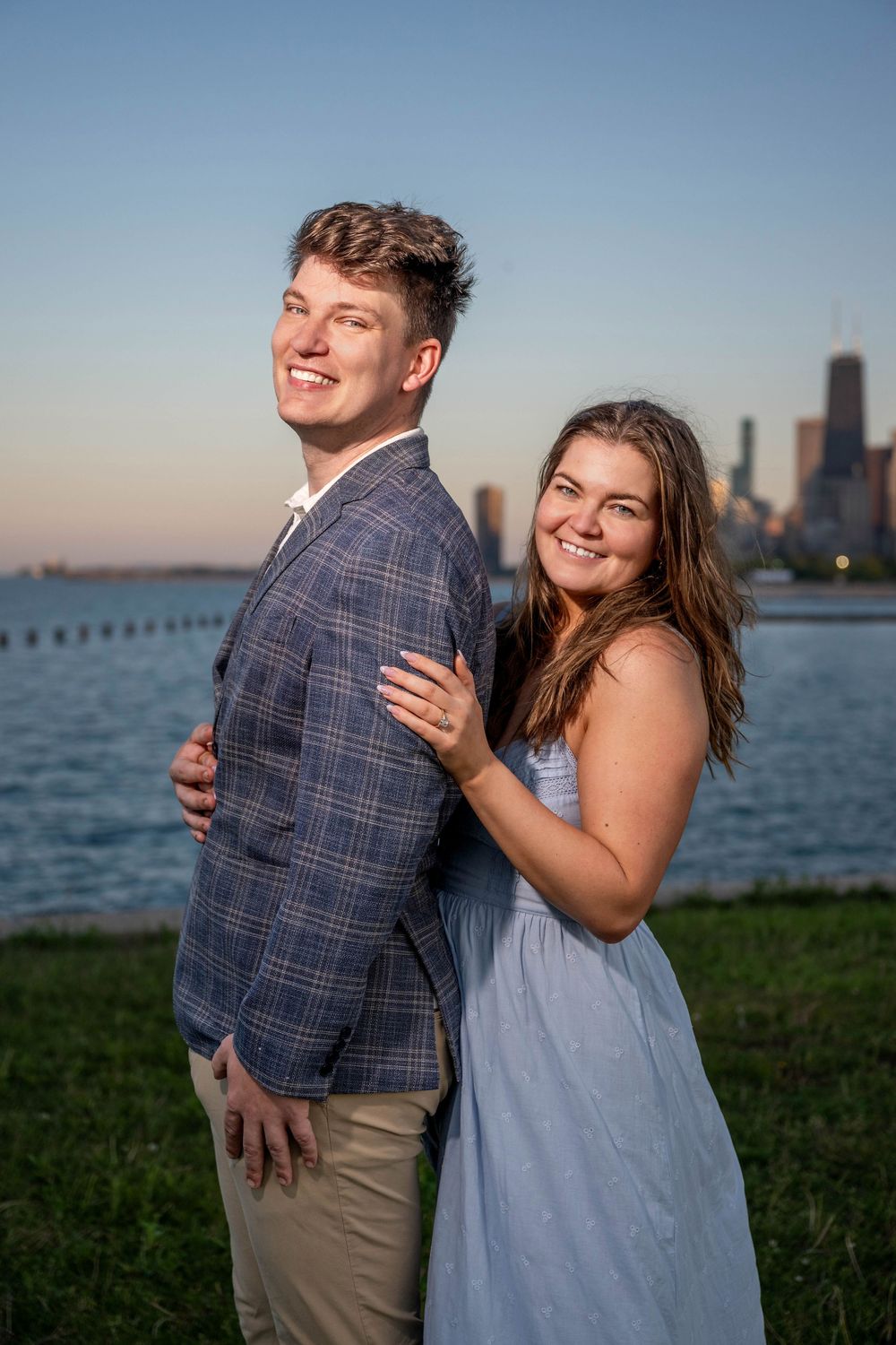 Smiling couple posing by Chicago lakefront at sunset, woman in blue dress embracing man in plaid blazer.