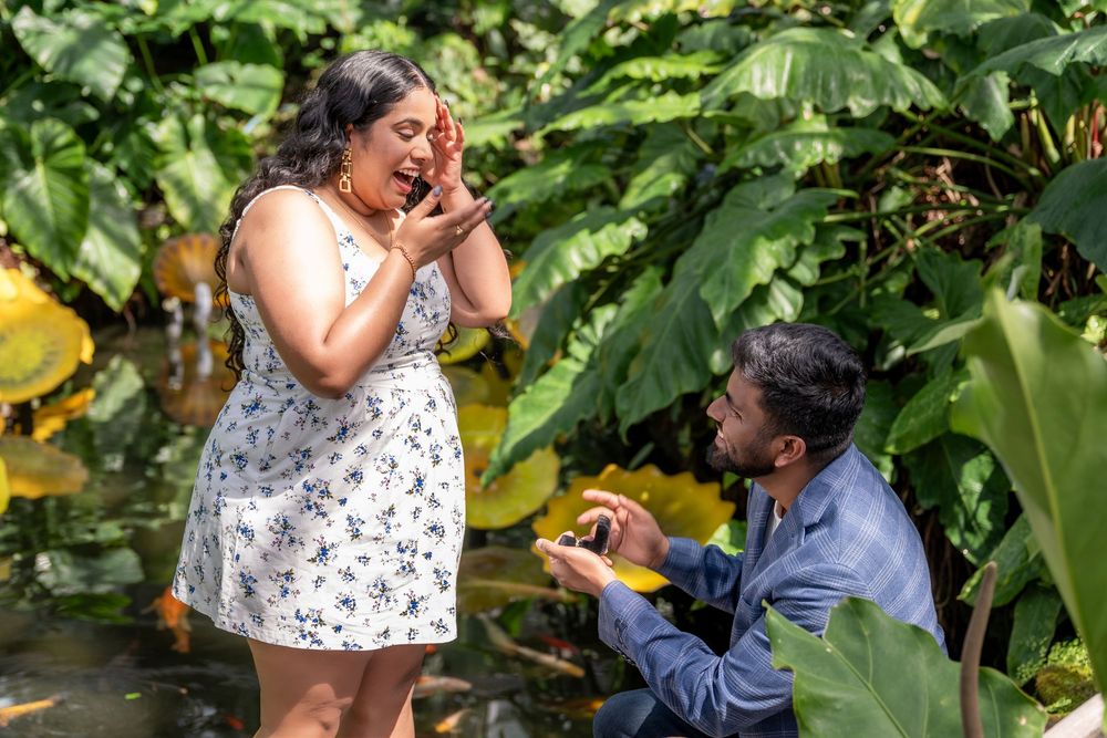 Man kneeling to propose to a surprised woman in a lush tropical garden setting.