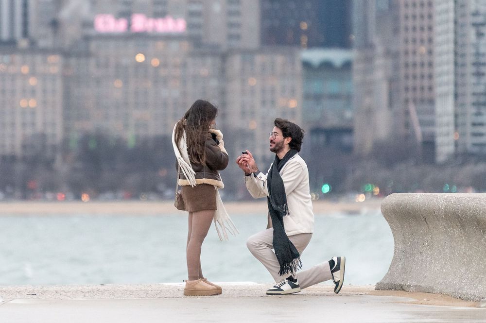 Man proposes to woman on one knee by Chicago lakefront with city skyline in background.