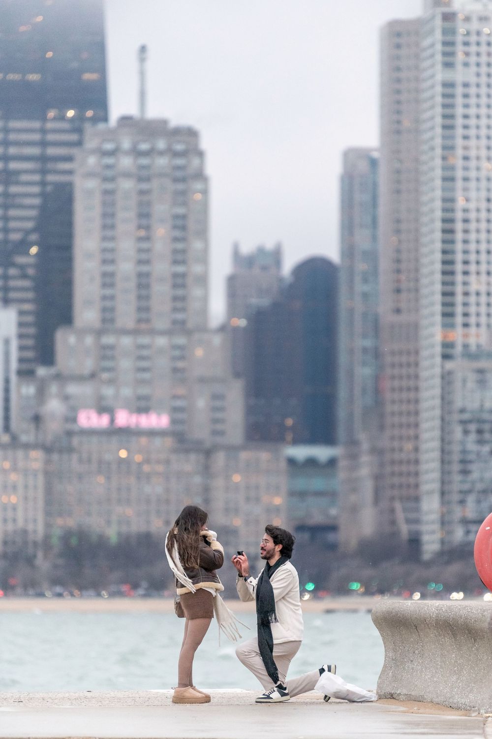Man proposes on one knee to woman by Chicago lakefront with city skyline in background.