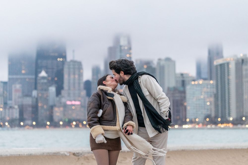 Couple kissing on a beach with a foggy city skyline in the background on a cold winter day.
