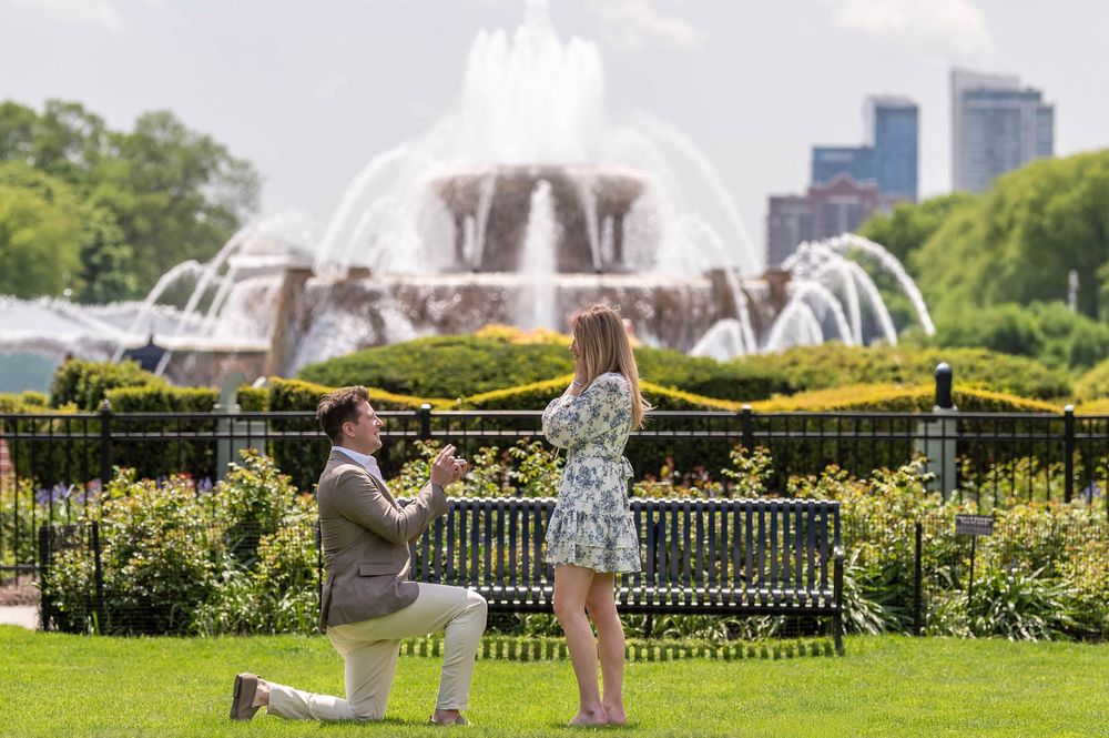 Man proposes on one knee to woman in front of Buckingham Fountain in Chicago's Grant Park on sunny day.
