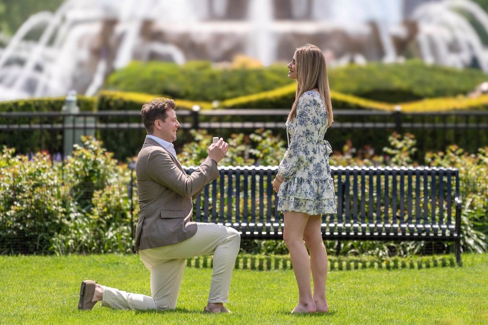 Man proposes to woman on one knee in a garden park with a fountain and green hedges in the background.