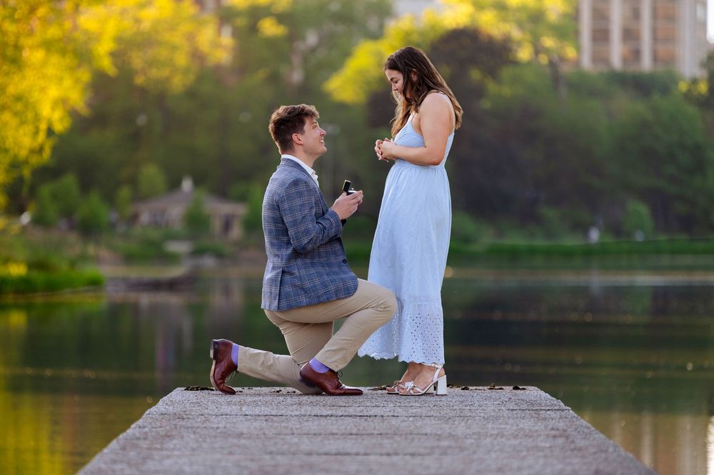 Man proposing to woman on a lakeside dock at sunset, surrounded by lush greenery in a romantic outdoor setting.