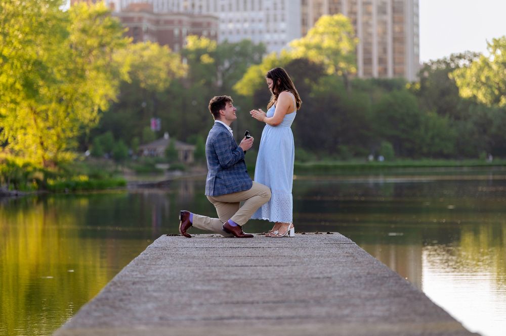 Man proposing to woman on a dock by a lake in a park, with lush green trees reflecting in the calm water.